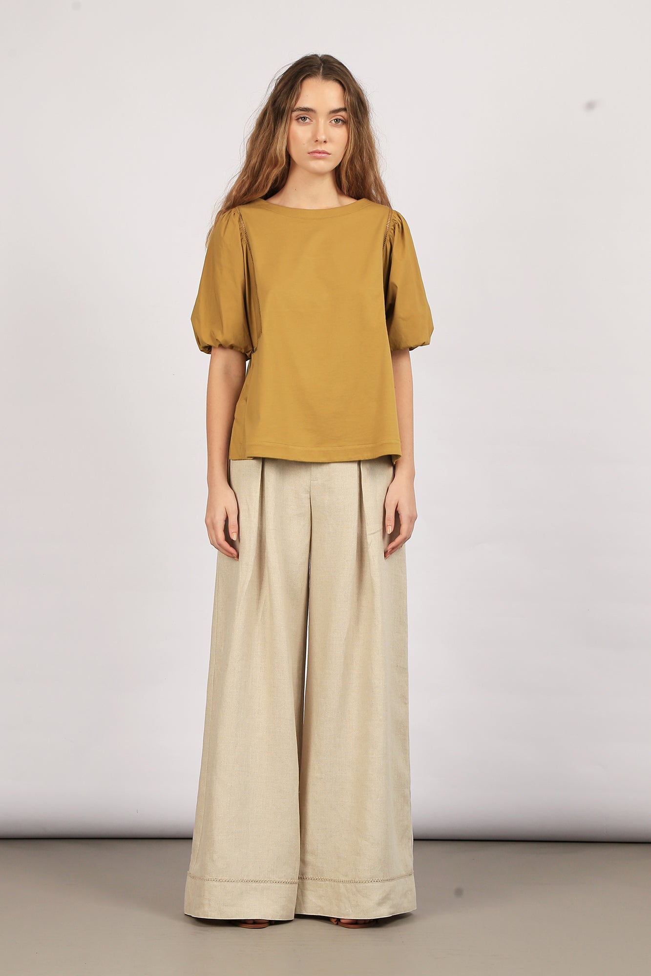 A model wearing an olive green Jamie Top with a round neckline and ¾ sleeves, paired with light-colored wide-leg pants.