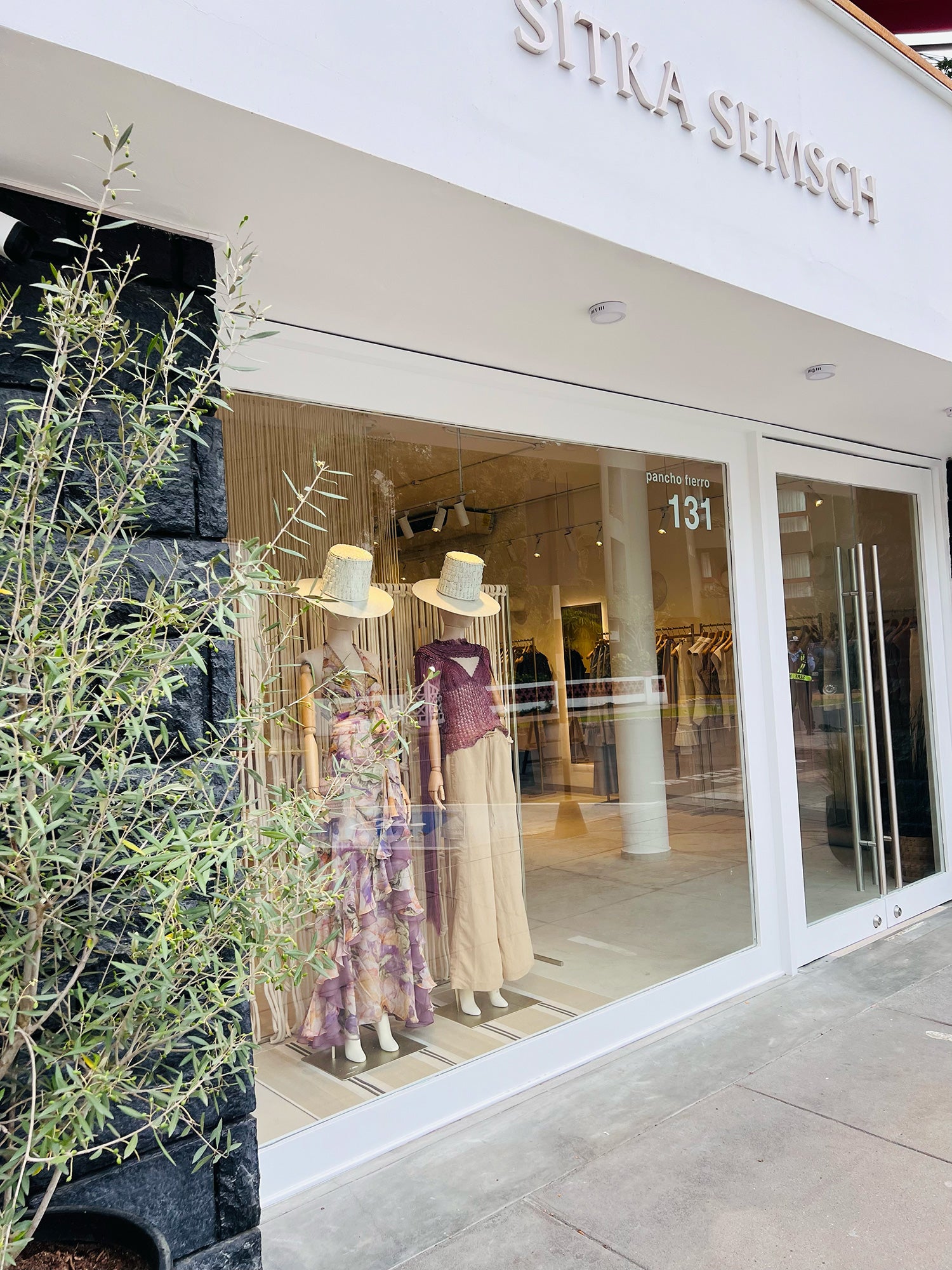 Daytime facade of the Sitka Semsch store in Lima, with the number 131 on the door. Two mannequins in the window display a patterned dress and wide-leg pants, both styled with straw hats.