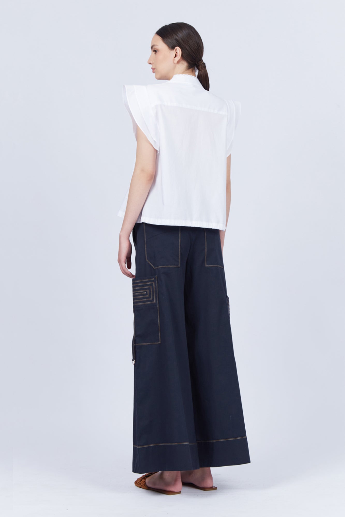 Pria Blouse in White – Sleeveless pima cotton blouse with V-neckline, Greca trim, tonal stitching, ruffle sleeves, and hem pullers.